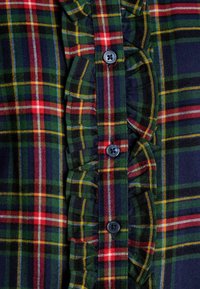 Plaid shirt featuring a mix of green, navy, red, and yellow colors, with a ruffled placket and button fastenings. Fabric is textured.