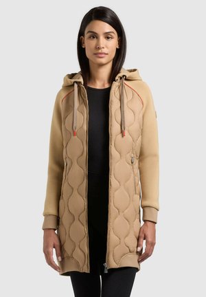 Nő bézs színű, kapucnis, steppelt dzsekiben, elöl cipzárral, fekete felsővel, egyszínű szürke háttér előtt állva.