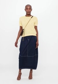 Gul tekstureret strikket kortærmet top, parret med en mørk denim maxi-nederdel med hvid syning og brune accessories.