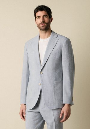 Man met een lichtblauw-wit gestreept colbert en bijpassende broek, gedragen over een wit overhemd, staand tegen een beige achtergrond.