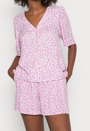 Vrouw draagt bijpassende roze en witte korte mouwen V-hals top en korte broek met bloemenprint, met handen in de zakken tegen een effen achtergrond.