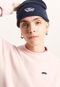 Gorro de canalé de color navy con el logo "Vans Off The Wall"; sudadera suave de color rosa con un pequeño bordado de una patineta; fondo neutro.