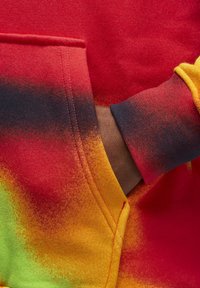 Main dans la poche avant d'un sweat à capuche coloré avec un tissu dégradé rouge, jaune, noir et vert.
