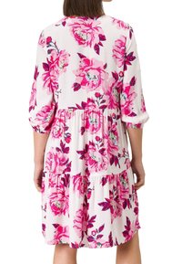 Femme portant une robe blanche mi-longue aux motifs floraux roses vifs et grands, avec des manches trois-quarts bouffantes, vue de dos.