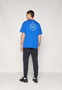 Blå bomulds t-shirt med korte ærmer, med et cirkulært hvidt logo med "CFC" på ryggen. Parret med sorte tapered sweatpants og sneakers.