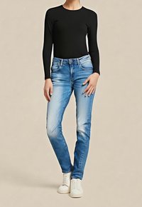 Persona vistiendo vaqueros ajustados de color azul claro, camisa negra de manga larga y zapatillas blancas, de pie contra un fondo liso beige.