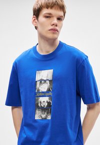 Μπλε βαμβακερή μπλούζα με κάθετη ασπρόμαυρη γραφική εκτύπωση που απεικονίζει χαρακτήρες σε στυλ anime και το κείμενο "Jujutsu Kaisen" σε κίτρινο χρώμα.