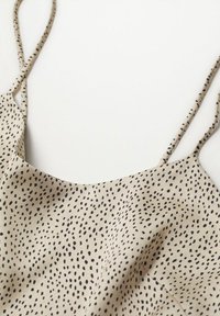 Haut beige à fines bretelles avec imprimé de points noirs irréguliers, tissu doux tombant sur un fond clair.