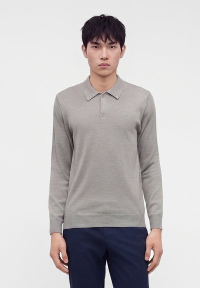 Jeune homme avec des cheveux noirs courts, portant un polo gris clair à manches longues et un pantalon bleu foncé, debout devant un fond blanc uni.