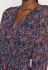 Květnaté šaty s tmavě modrým pozadím, na kterém jsou malé červené a bílé květy, véčkový výstřih a nařasené detaily na živůtku.