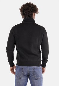 Maglione nero a coste con colletto alto; presenta una vestibilità aderente, maniche lunghe e una superficie testurizzata; abbinato a jeans in denim blu.
