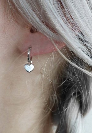 Silberne Creolen mit einem kleinen Herz-Anhänger mit strukturierten Oberflächen, sichtbar am Ohr einer Person mit hellem, welligem Haar.