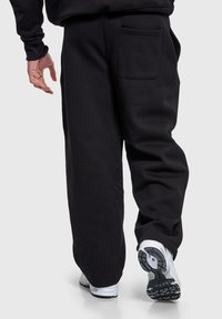 Pantalon de survêtement noir en tissu doux, avec une poche arrière et une coupe décontractée, associé à des baskets de sport blanches aux semelles texturées.