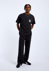 Black cotton t-shirt with short sleeves and a graphic design on the chest, paired with black wide-leg jeans and black sneakers.