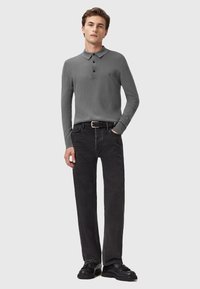 Ung mand iført en grå langærmet polo, sort bælte, sorte lige ben jeans og sorte loafers med kvaster, står med den ene hånd i lommen.