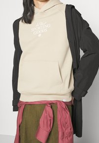 Béžová mikina s kapucí a přední kapsou s bílým logem, vrstvená pod černou vnější bundou, doplněná růžovou košilí svázanou přes zelenou sukni.