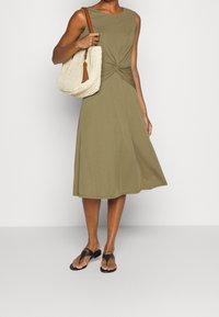 Zielonooliwkowa sukienka bez rękawów z skręconym detalem z przodu, luźna spódnica, w towarzystwie plecionej beżowej torby z frędzlami. Czarne klapki.