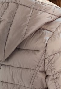Lysebrun quiltet jakke med hætte, synlige syninger og polstrede sektioner til varme, båret på ryggen og skulderområdet.
