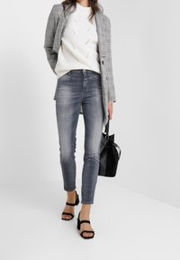 Blazer de cuadros gris sobre un suéter blanco texturizado, acompañado de jeans ajustados de mezclilla clara y sandalias negras de tacón bloque. Bolso negro en una mano.