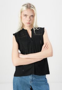 Bluză neagră fără mâneci, cu guler, realizată dintr-un material texturat cu buline. Are volane la umeri, închidere cu nasturi și detalii delicate de feliere.