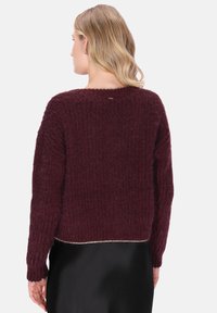 Kvinde med blond hår iført en struktureret mørk burgunder striktrøje og sort nederdel, vist fra ryggen mod en hvid baggrund.