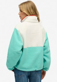 Veste en polaire aqua et crème, avec un col montant aux accents rouges, coupe oversize et tissu texturé.