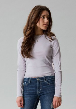 Ung kvinde med langt brunt hår iført en lysegrå/lilla bluse med lange ærmer og blå jeans, som kigger mod højre mod en ensfarvet baggrund.