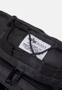 Borsa nera in nylon con chiusura a cerniera, cordino regolabile e etichetta bianca con testo che riporta informazioni sul marchio e sulla produzione.