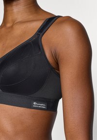 Melns sporta krūšturis ar regulējamām lencītēm, izgatavots no gludas auduma. Ir polsterēts dizains, šūšanas detaļas un "Champion Shock Absorber" uzlīme.