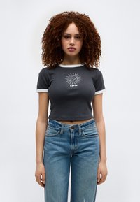 Νεαρή γυναίκα με σγουρά μαλλιά φοράει μαύρο κροπ τοπ με σχέδιο ήλιου και λογότυπο Levi’s, συνδυασμένο με μπλε ψηλόμεσα τζιν.