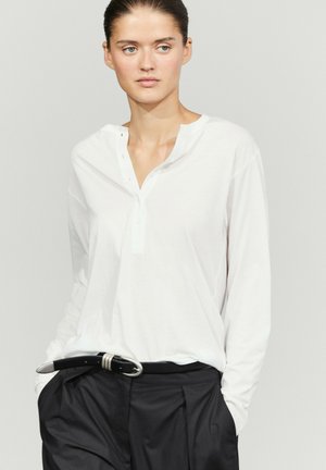 Woman with dark hair tied back wearing a loose white long-sleeve shirt and black pants with a black belt, hands in pockets, looking to the side.