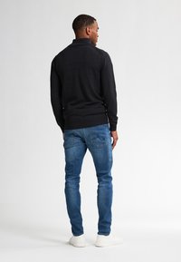 Schwarzer, langärmeliger Pullover mit Kragen, kombiniert mit blauen Jeans. Weiße Sneakers runden das lässige Outfit ab. Modell von hinten positioniert.