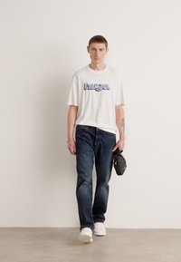 White cotton t-shirt with blue "hugo" logo, paired with dark denim jeans and white sneakers, holding a small black bag. Plain background.
