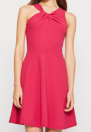 Robe rose vif sans manches avec un col halter torsadé et une jupe évasée portée par une femme sur un fond uni.