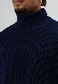 Man draagt een donkerblauwe geribde koltrui, close-up die de textuur en het kraagdetail toont.