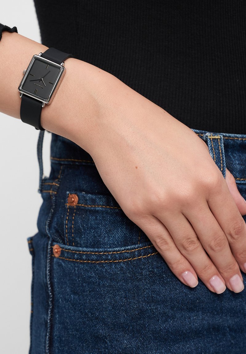 Quadratische Analog-Uhr mit schwarzem Ziffernblatt, silbernem Metallrahmen und schwarzem Silikonarmband, getragen an einem Handgelenk gegen dunklen Jeansstoff.