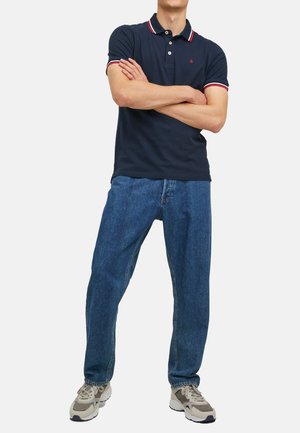 Marineblå polo-shirt med rød og hvid stribet krave, parret med løstsiddende blå denim jeans og grå sneakers.