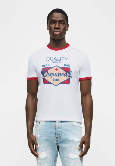 Άνδρας που φοράει λευκό T-shirt με κόκκινο γιακά και άκρα μανικιών, τυπωμένο με τη φράση «QUALITY tested by Dean Dan Dsquared2 Bros» και ανοιχτό μπλε τζιν με σκισίματα.