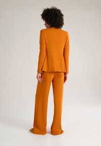 Costume orange en deux pièces comprenant un blazer ajusté avec une coupe structurée et un pantalon à jambes larges, fabriqué à partir d'un tissu lisse avec une finition mate.