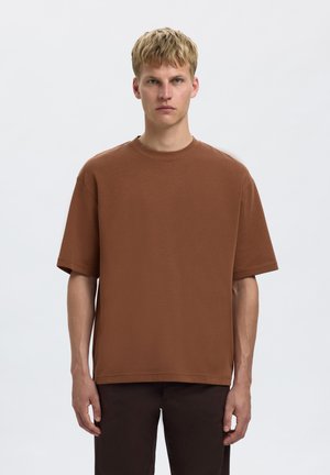 Jeune homme aux cheveux blonds courts portant une chemise marron ample à manches courtes et un pantalon foncé, debout devant un fond blanc uni.
