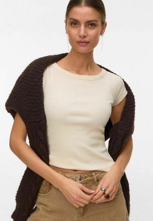 Kvinde iført en cremefarvet kortærmet top, brun tekstureret strik lagt over skuldrene, beige bukser, hængende øreringe og to ringe på venstre hånd.