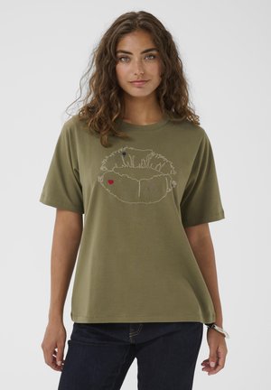 Jonge vrouw met golvend bruin haar draagt een olijfgroene t-shirt met een grote witte geborduurde lipontwerp met kleine rode en blauwe details.