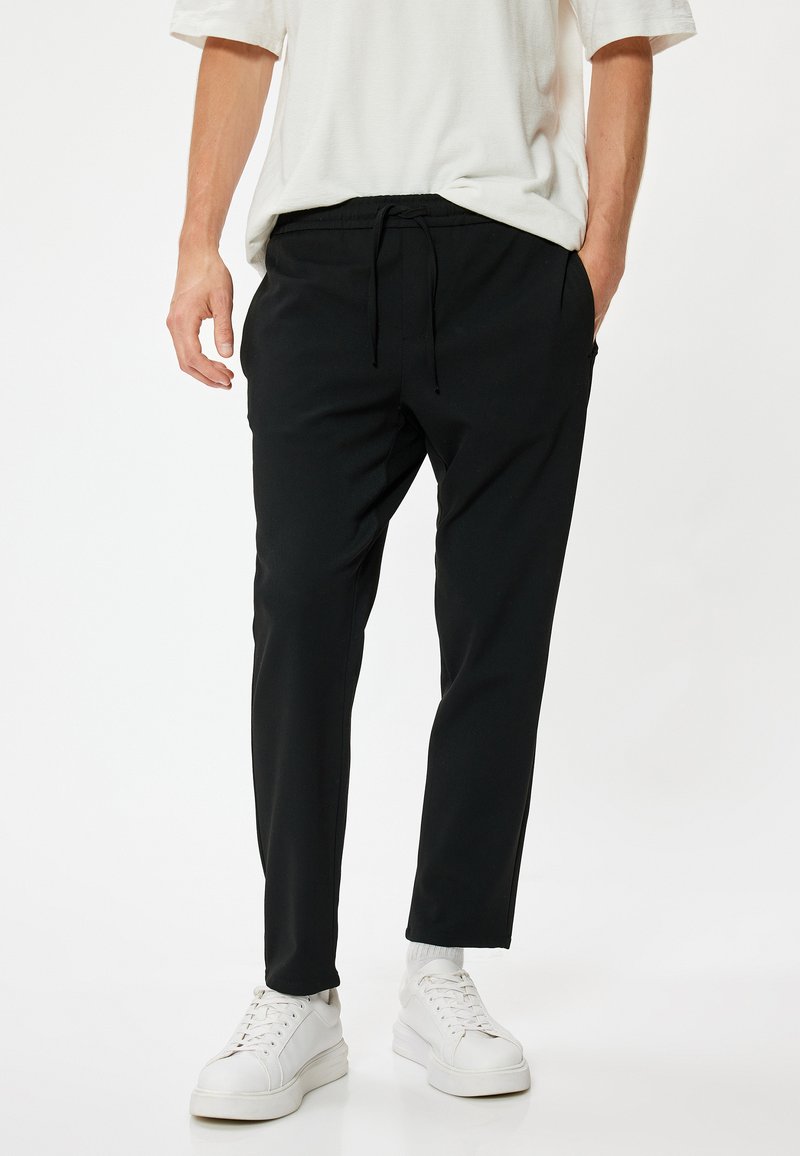 Zwarte taps toelopende broek met een elastische tailleband en trekkoord, voorzien van zijzakken, gecombineerd met witte sneakers. Glad textiel.