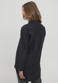 Black long-sleeve shirt with a buttoned collar, featuring a loose fit and a curved hem. Smooth texture, minimalistic design without patterns.