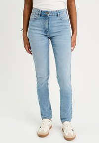 Kvinde iført lyseblå slim-fit jeans og hvide sneakers med beige og sorte nuancer, stående mod en ensfarvet hvid baggrund.