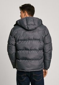 Tamno siva puffer jakna s kvadratnim uzorkom i kapuljačom, s glatkom, blago sjajnom teksturom. Nosena preko plavih traperica.