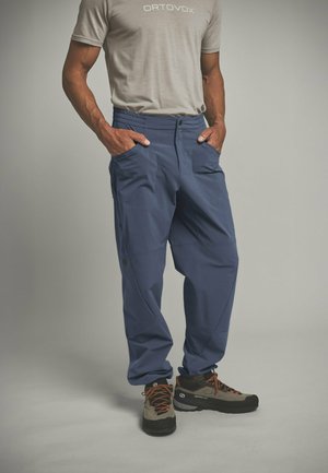 Mann trägt ein hellgraues Ortovox-T-Shirt, blaue Wanderhose und graue Wanderschuhe mit orangefarbenen Schnürsenkeln, steht mit den Händen in den Taschen.