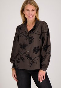 Glimlachende vrouw met lang blond haar, die een donkerbruine blouse met knopen en zwarte bloemenpatronen draagt, en een zwarte broek, staand tegen een witte achtergrond.