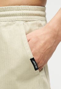 Το δεξί χέρι ακουμπά μέσα στην μπροστινή τσέπη ενός μπεζ παντελονιού με ελαστική μέση, που φέρει μια μικρή υφασμάτινη ταμπέλα "Santa Cruz".