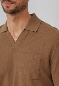 Brązowy pulower krótki z rękawami, o prążkowanej teksturze, z dekoltem w kształcie litery V oraz naszywaną kieszenią na lewej piersi. Wykonany z miękkiego dzianiny.